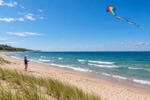 A person flying a colorful diamond kite with a long rainbow tail above a sandy beach.