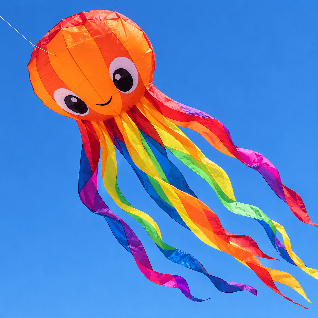 A smiling orange octopus kite with long rainbow ribbon tentacles flying against a blue sky.